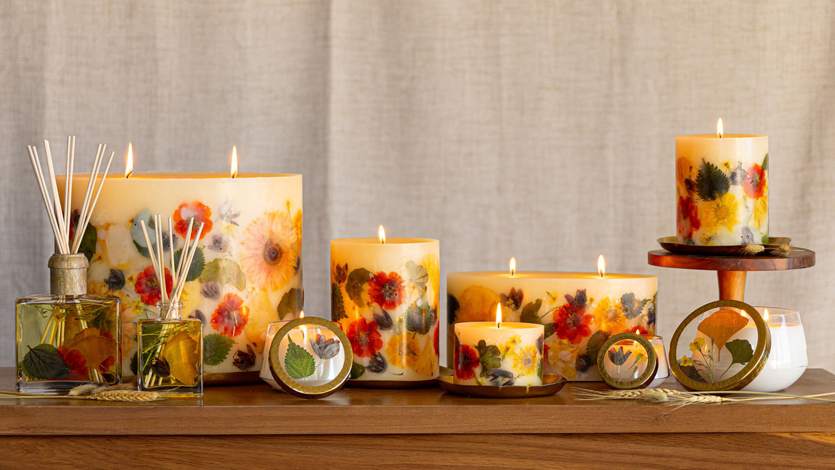 A collection of decorative candles and reed diffusers featuring colorful pressed floral designs in red, orange, yellow, and green, displayed on a wooden shelf against a neutral linen background.