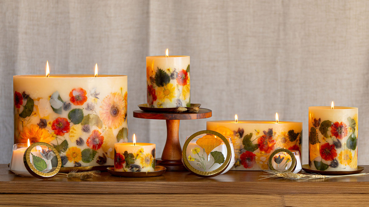 A collection of decorative pillar candles and small tins with pressed floral designs, displayed on a wooden surface against a neutral linen backdrop.